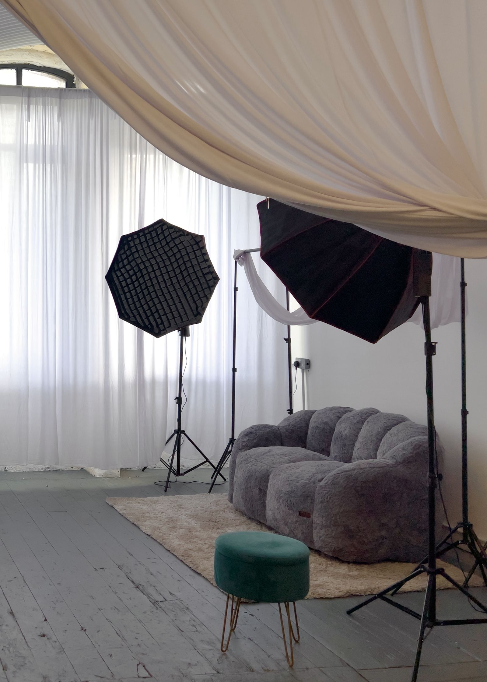 A grey scalloped sofa and green velvet stool under a draped ceiling, with softboxes